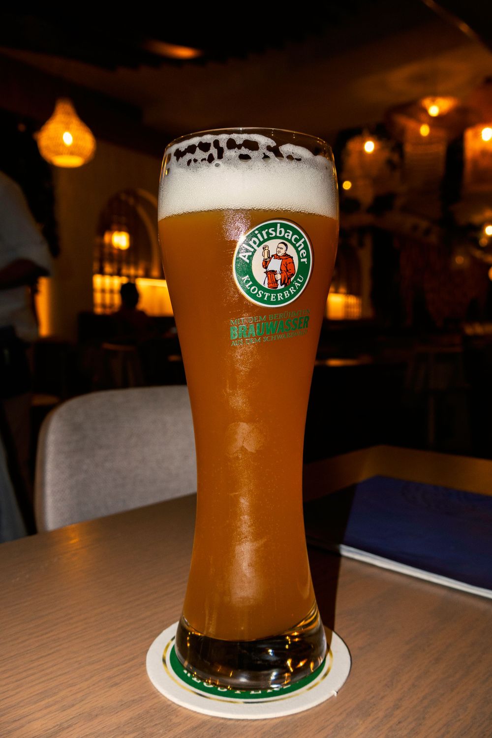 A tall glass of golden Alpirsbacher wheat beer with frothy head sits on a coaster in a dimly lit bar setting.
