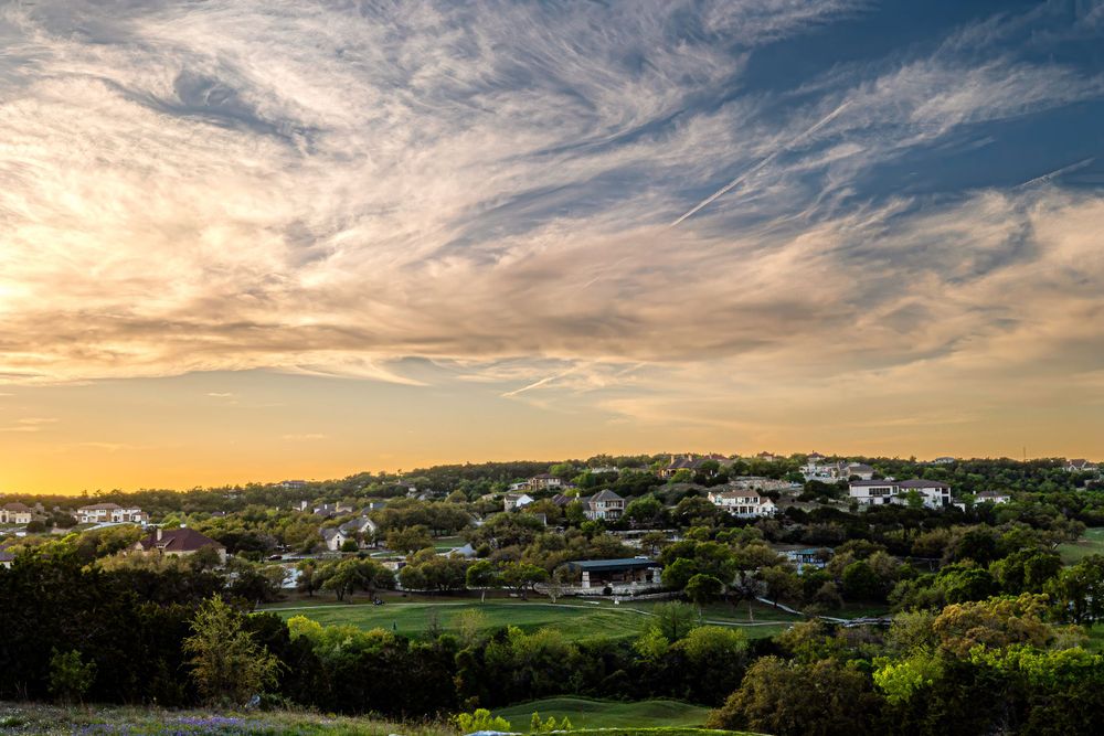 Scenic sunset view over a coastal town with white buildings nestled among green hills under dramatic wispy clouds.