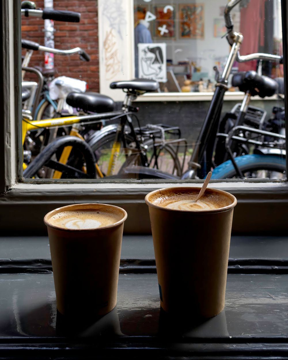 Two cups of coffee with latte art sit on a windowsill overlooking parked bicycles through the glass window.