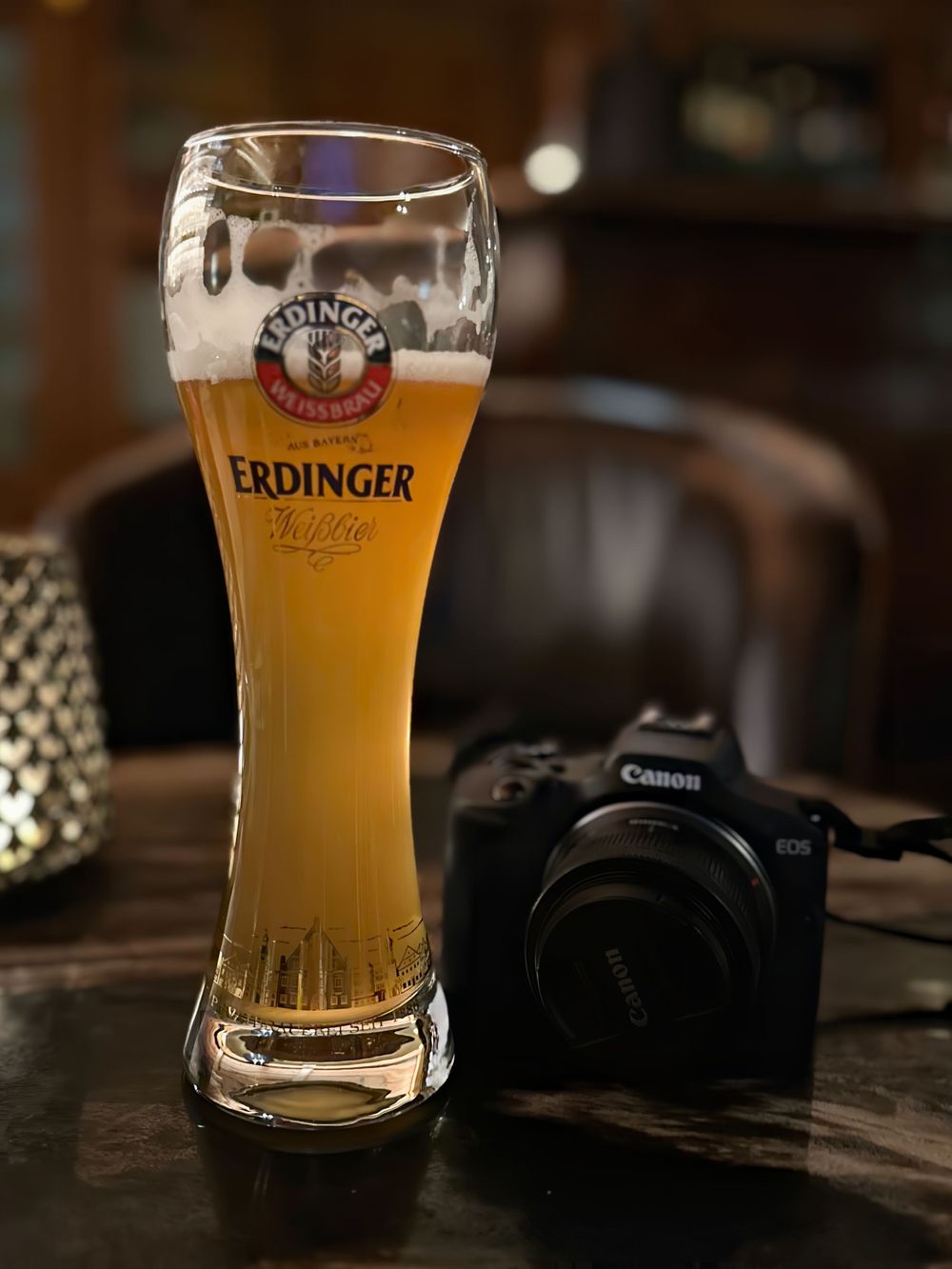 A glass of golden Erdinger wheat beer sits on a wooden table next to a Canon DSLR camera in dim lighting.