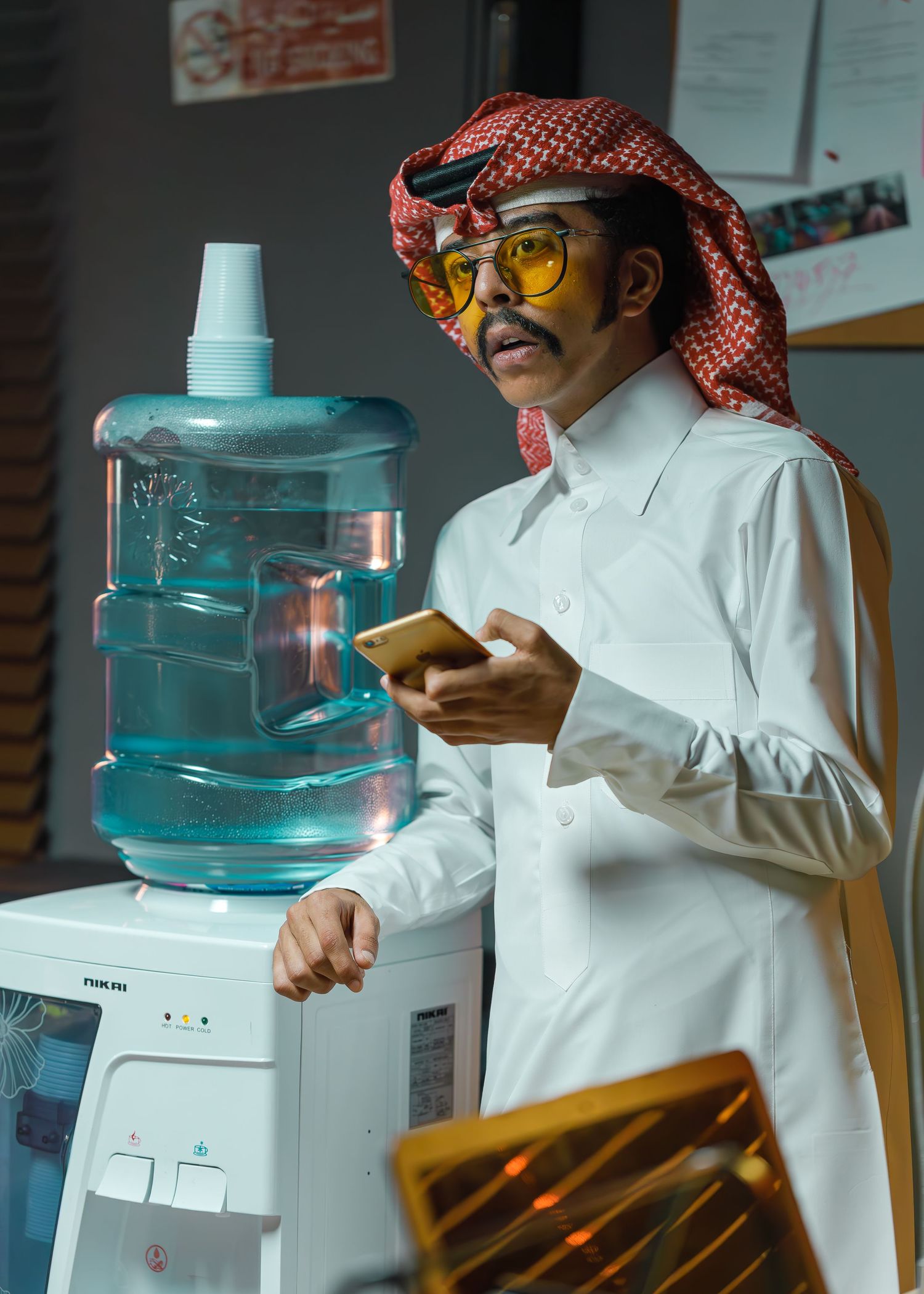 Person in traditional Middle Eastern dress and yellow glasses checks phone while standing next to a water cooler in an office.