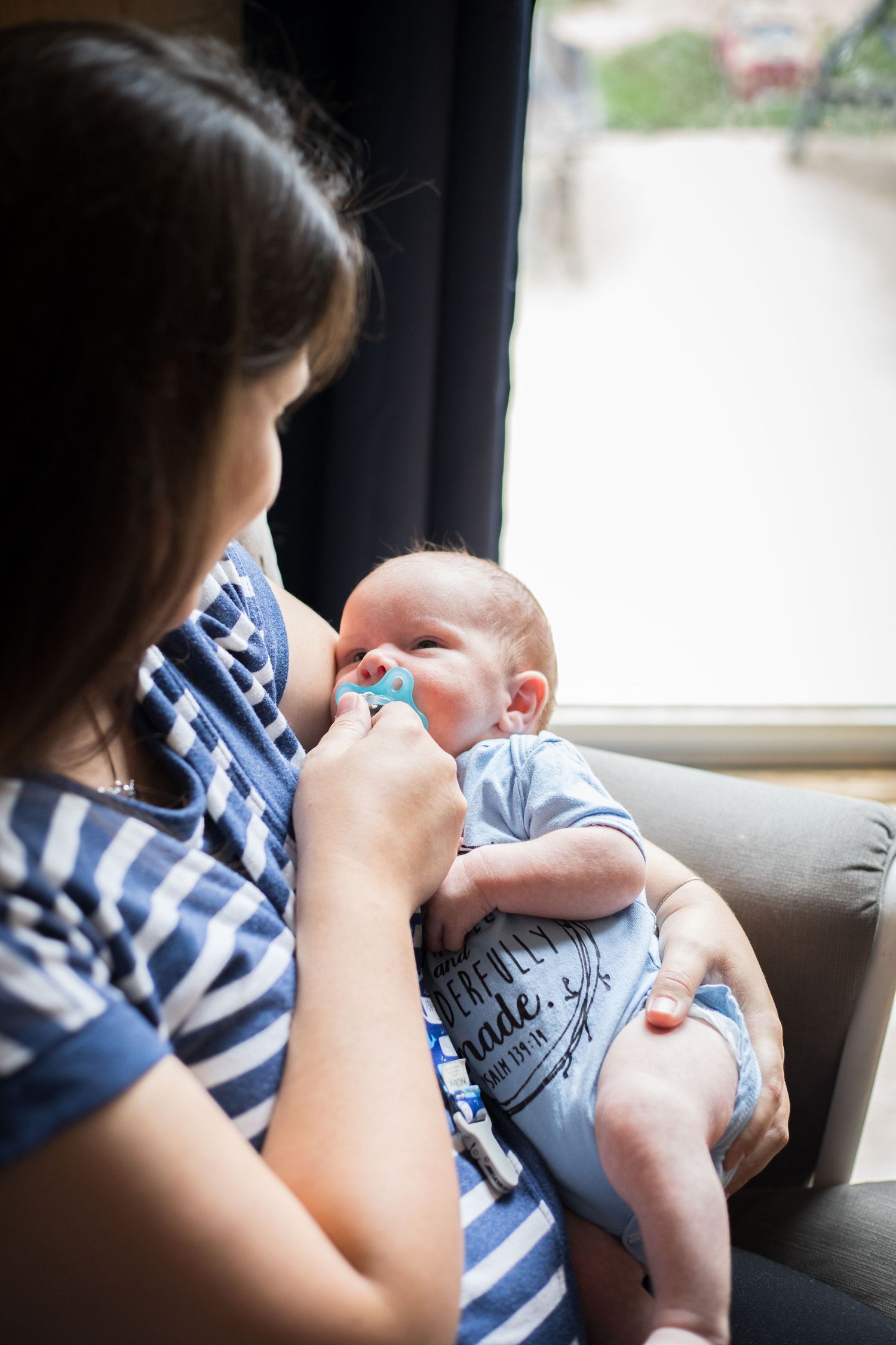 Mother holds newborn baby with pacifier while sitting by a window indoors.