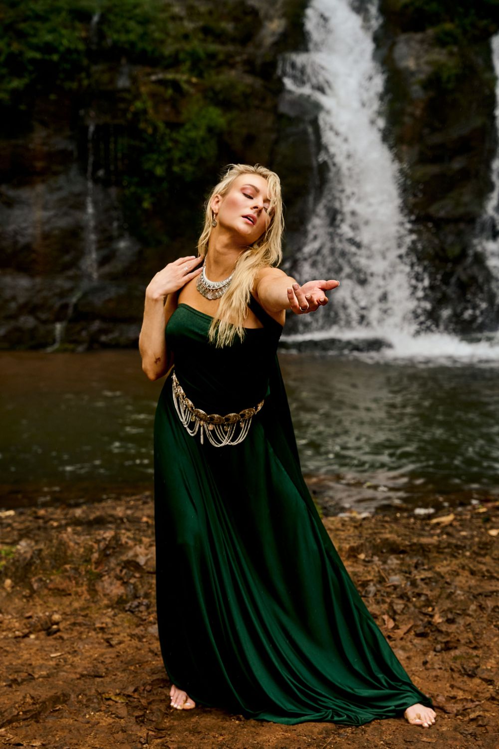 A person in a flowing emerald green dress strikes a graceful dance pose against a scenic waterfall backdrop.