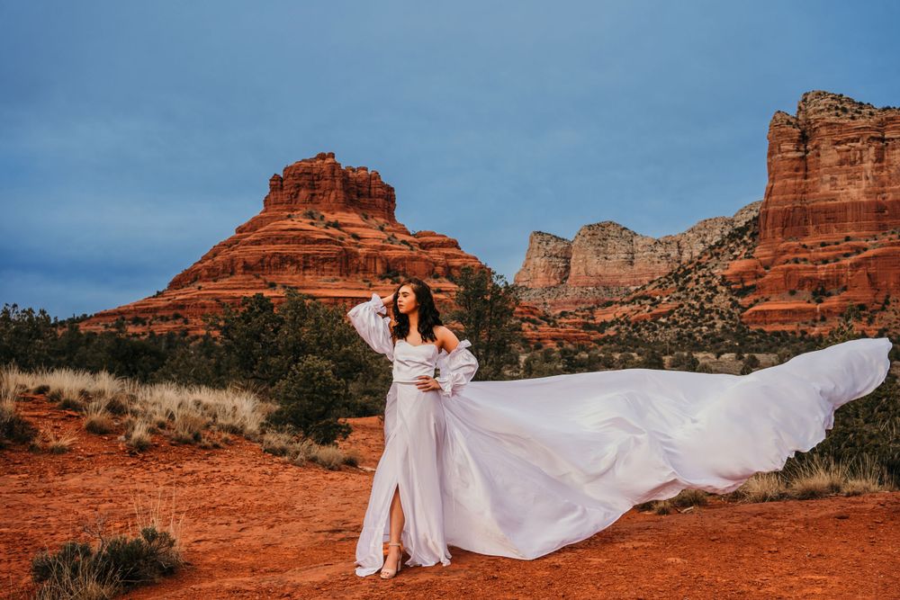Flowing white wedding dress billows in the wind against dramatic desert cliffs and blue sky.