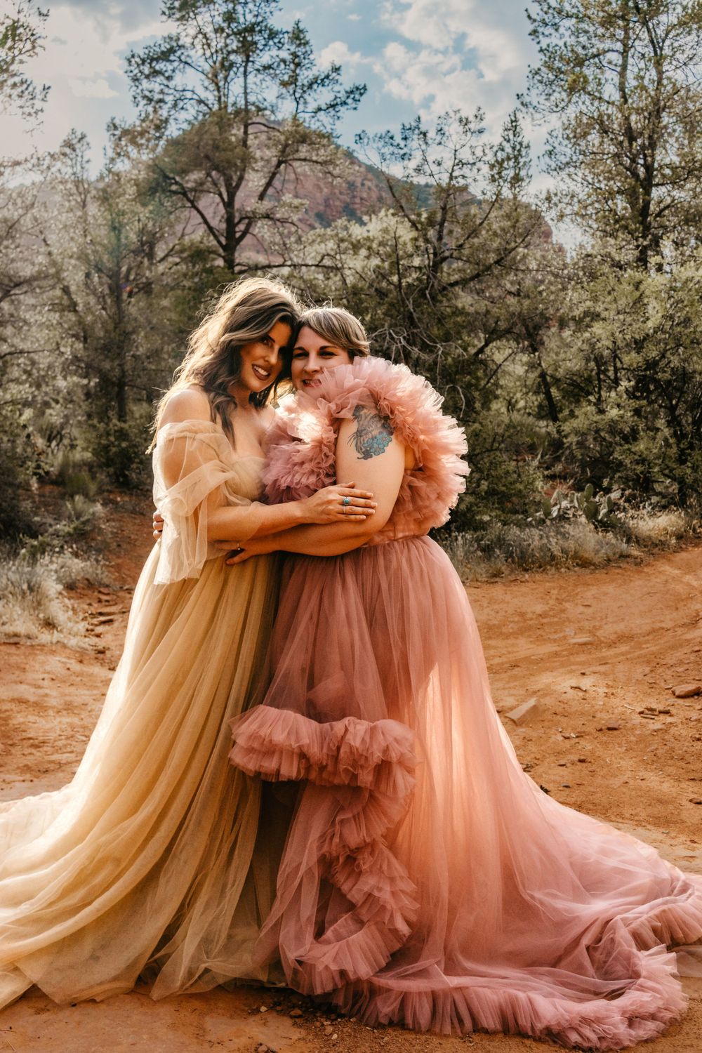 Two figures embrace in flowing peach and pink tulle gowns against Sedona's red rock landscape.