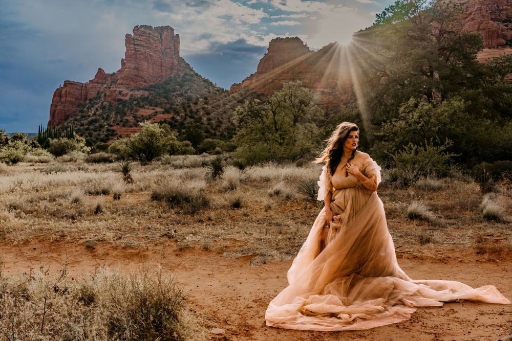 Elegant coral dress billows in desert breeze with dramatic red rock formations and sunburst in background.