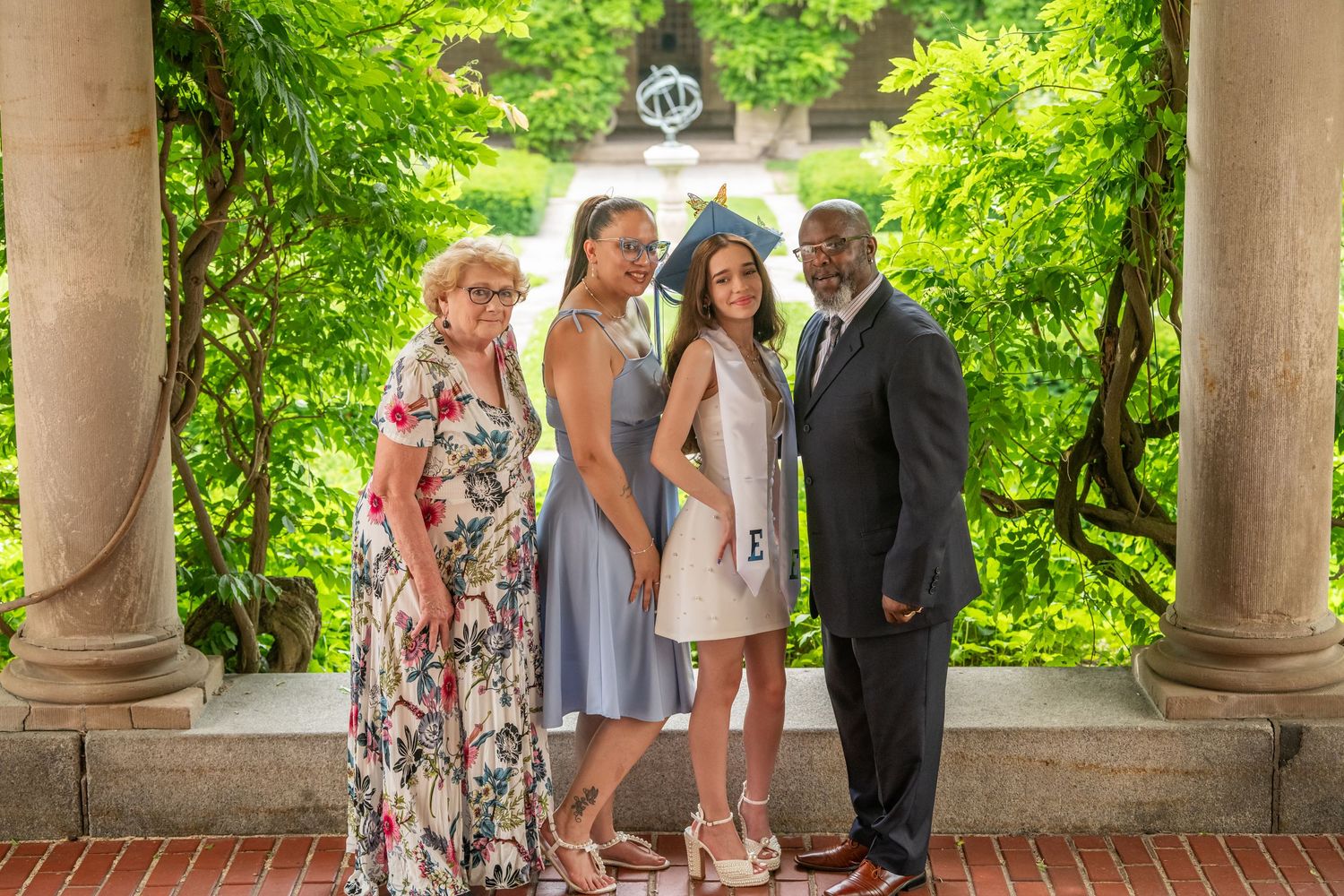 Family of four poses together under garden archway with lush greenery at outdoor graduation celebration.