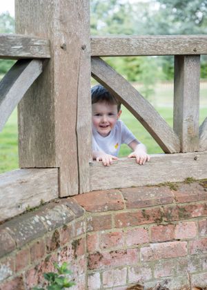 Young smiling boy peeking through weathered wooden gate and brick wall in garden.