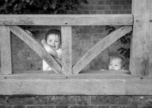 Black and white photo of two young children peeking through a rustic wooden fence.
