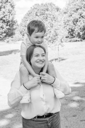 Black and white portrait of smiling mother carrying young child on her shoulders outdoors.