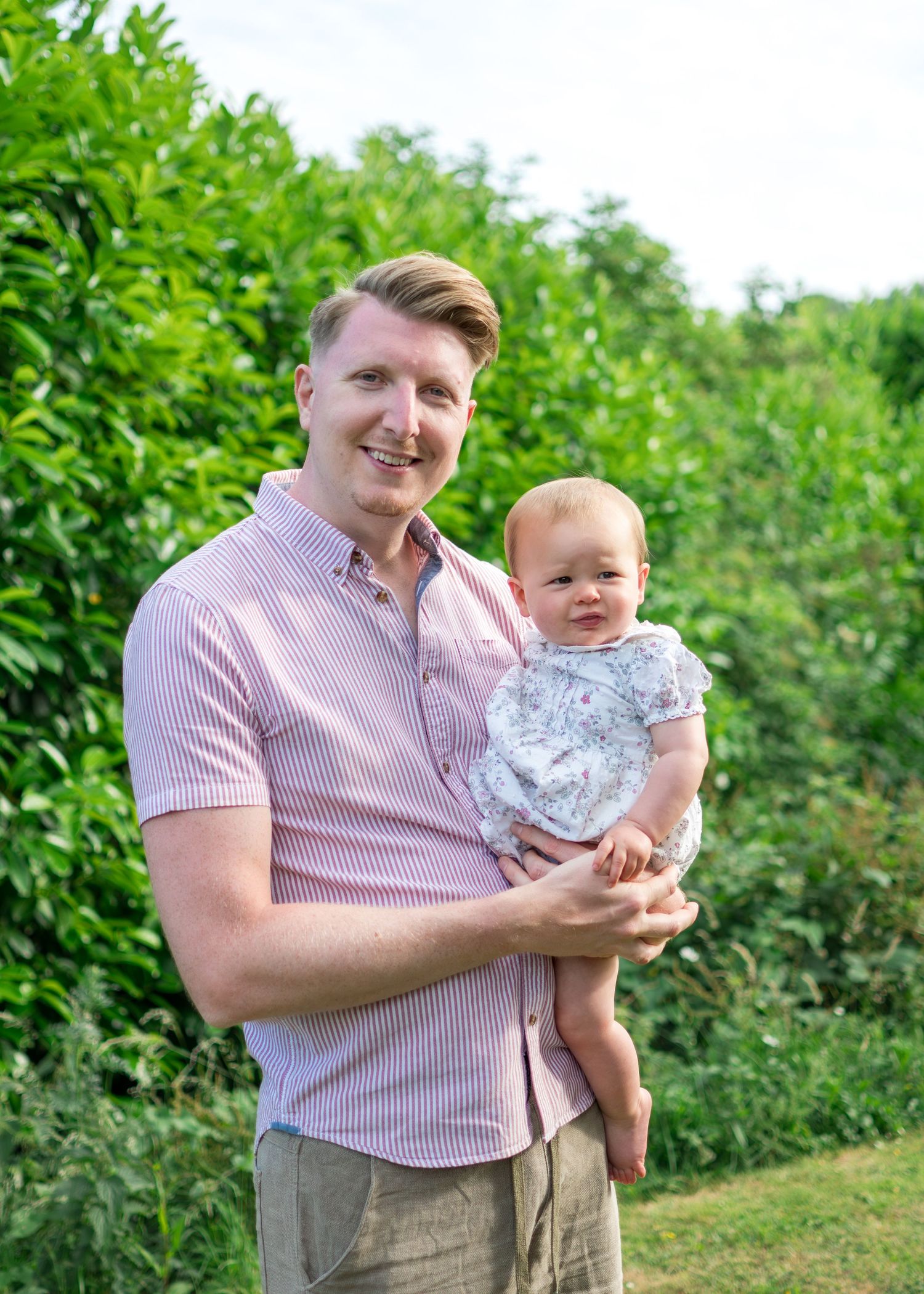Smiling man in pink shirt holding a baby outdoors in a green garden.