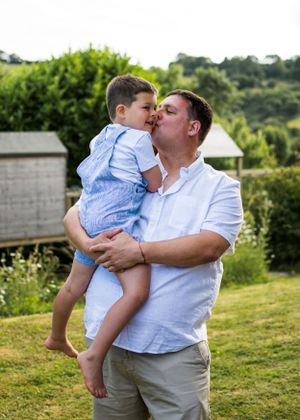 A father lovingly kisses his young son while holding him in a lush green garden on a sunny day.