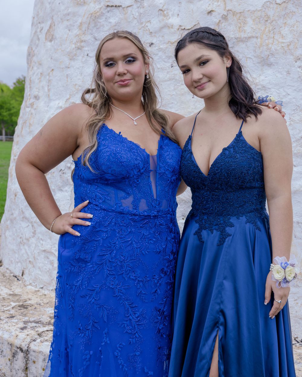 Two people in coordinating blue formal dresses pose together against a white stone wall.