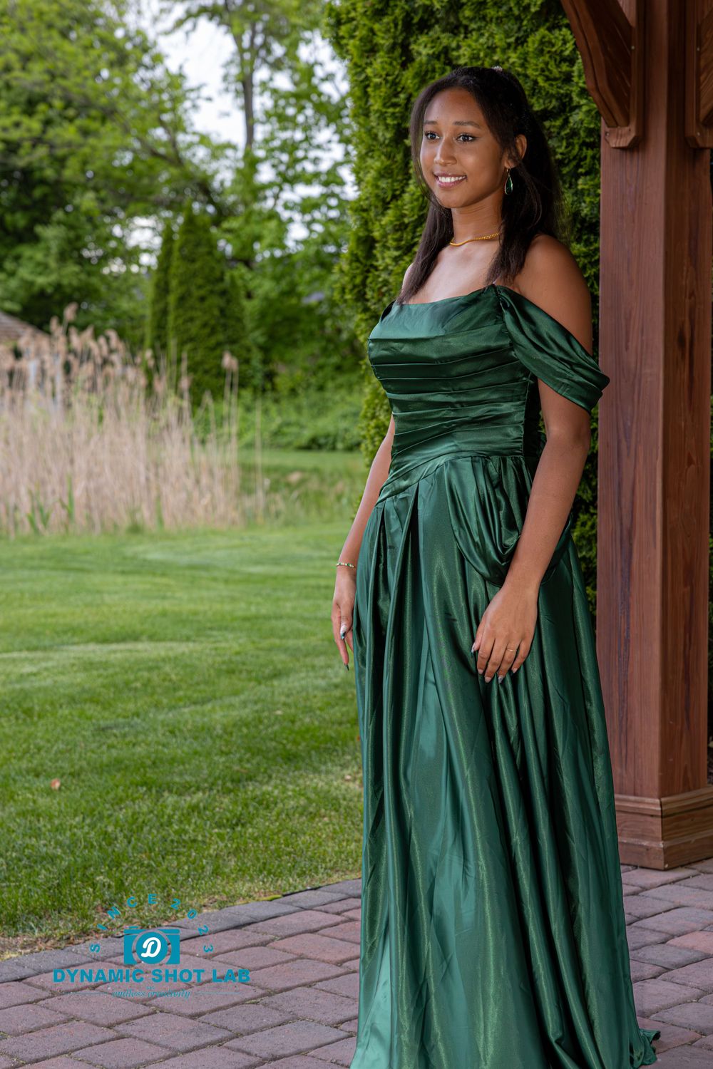 Person wearing an elegant emerald green formal dress poses in front of a wooden gazebo.