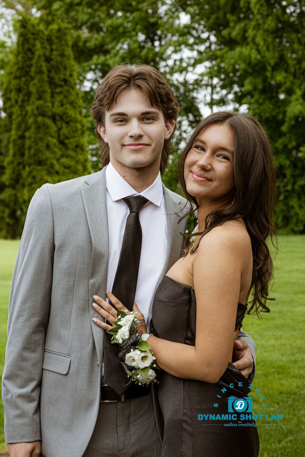 A couple in formal attire poses together on a grassy lawn during a special event.