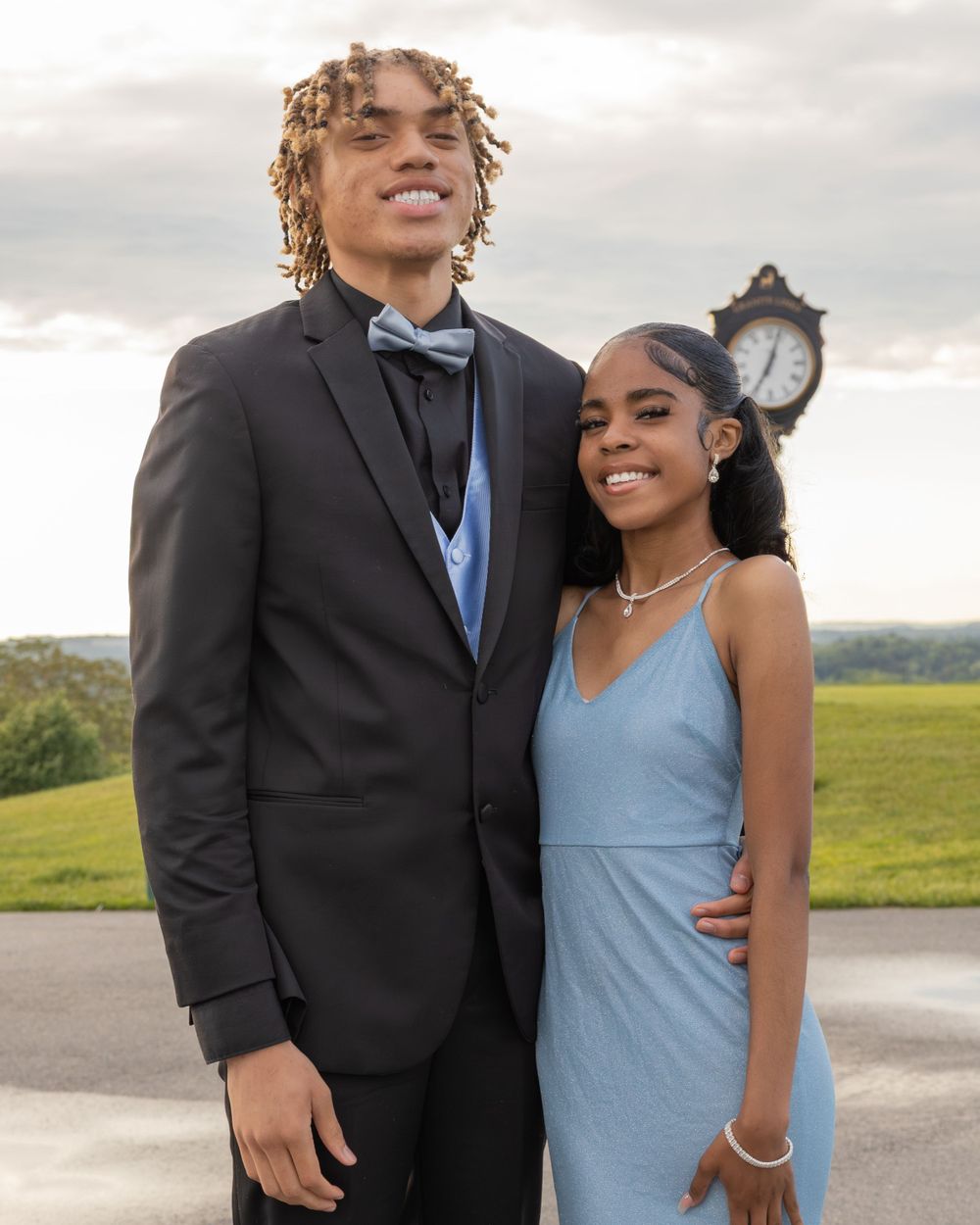 Formal couple poses together outdoors, one in black suit and one in light blue dress.