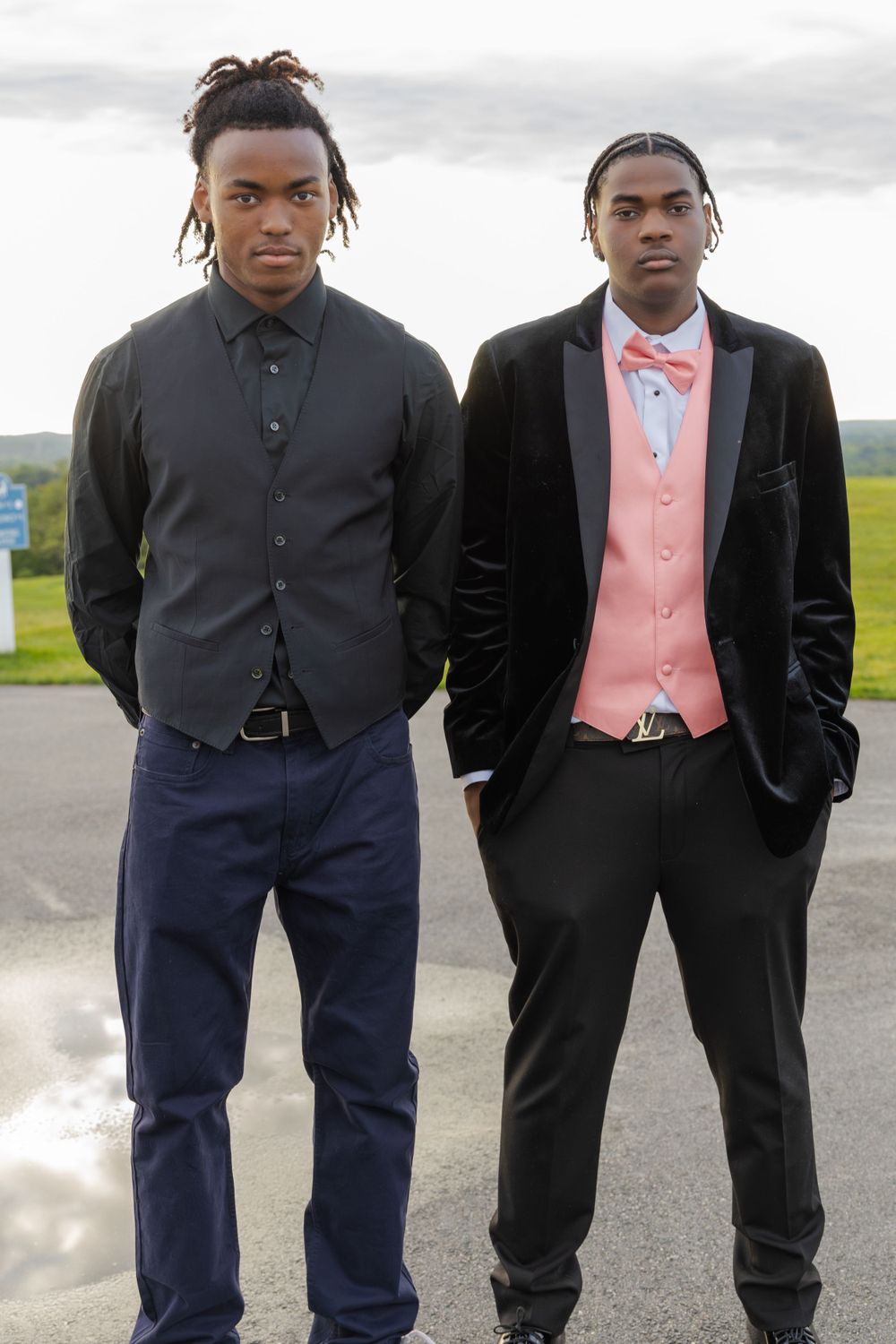 Two people in black formal suits stand together on paved path.