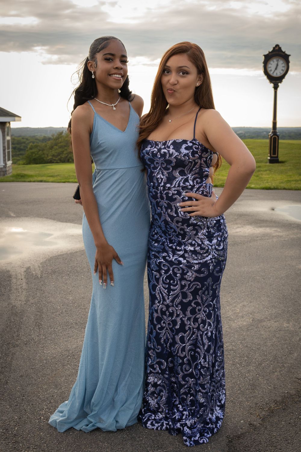 Two people in formal attire pose together on paved path with cloudy sky background.