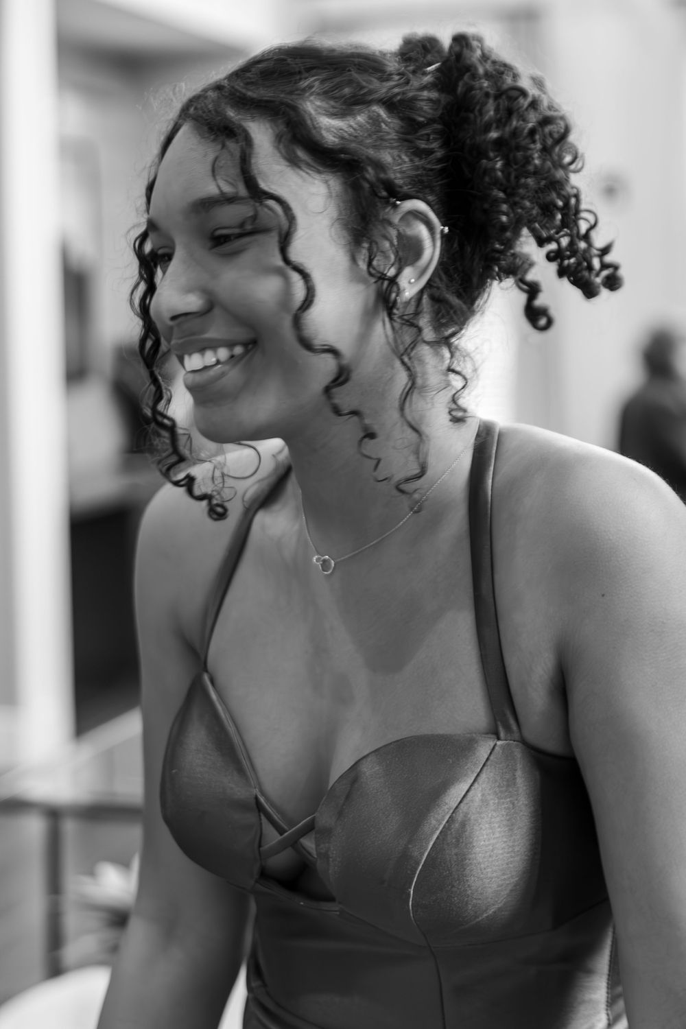 Black and white portrait of a smiling young woman with curly hair and a neck tattoo wearing a metallic dress.