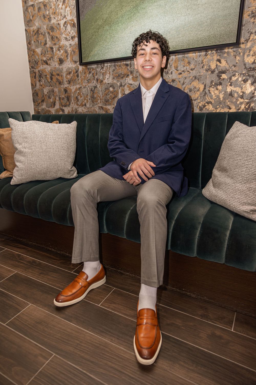 Young man in navy blazer and tan loafers sits smiling on a green velvet bench.