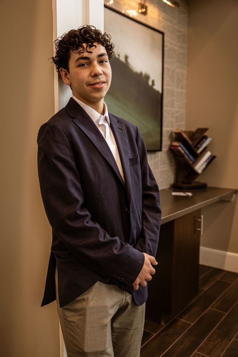 Young man with curly hair in navy blazer and khakis poses confidently in an elegant interior setting.