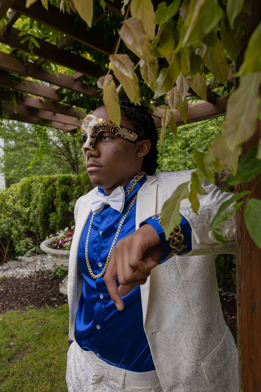 Someone wearing a blue vest and pearl necklace poses under a garden pergola.