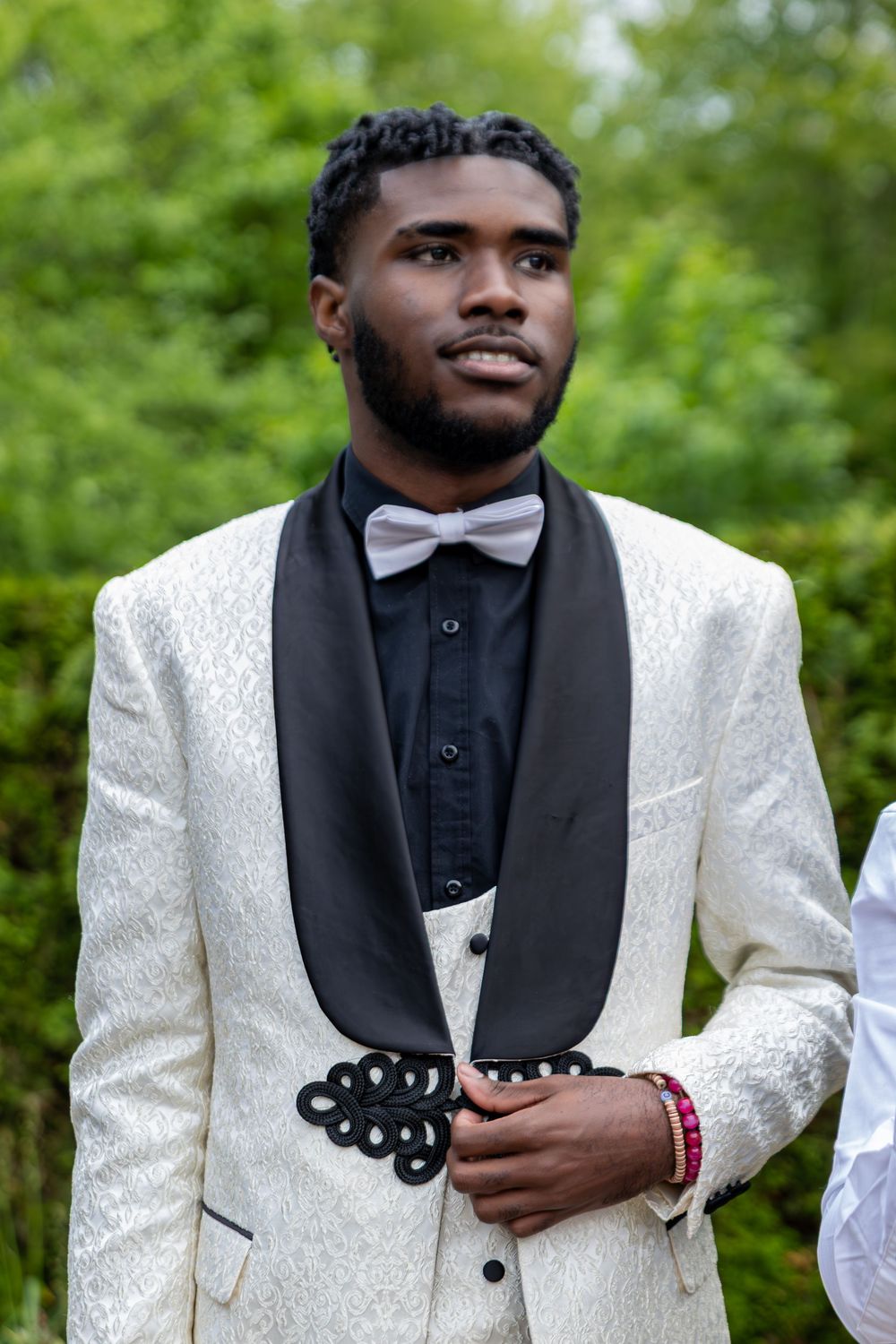A person wearing a white tuxedo jacket with black bow tie smiles in an outdoor setting.