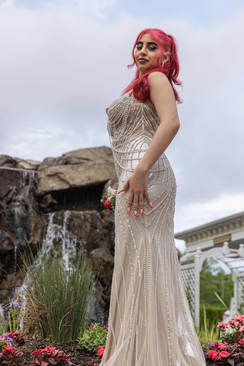 A silver sequined dress sparkles against a garden backdrop with a waterfall and pink flowers.