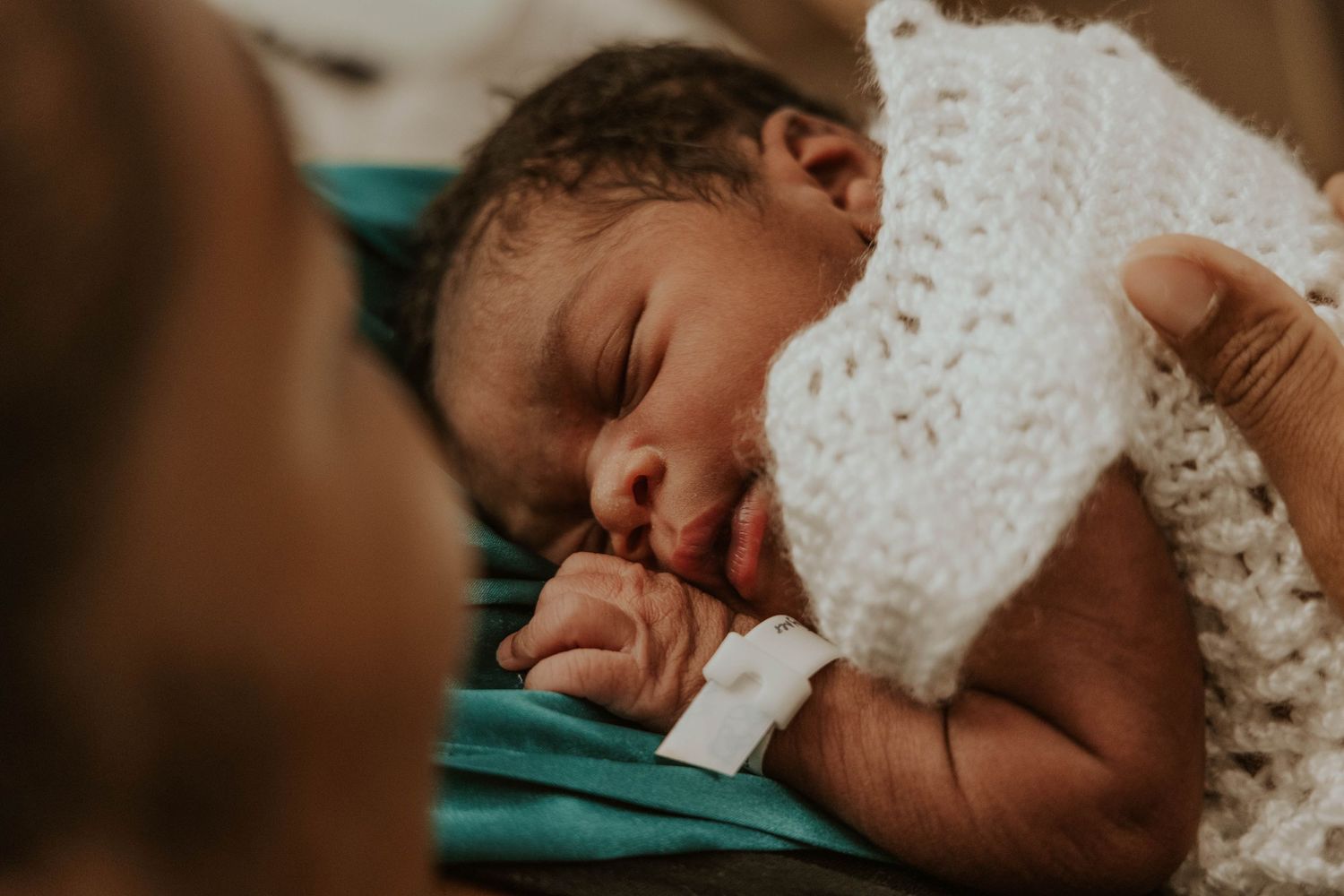 Close up of peaceful sleeping newborn wrapped in white knit blanket on teal background.