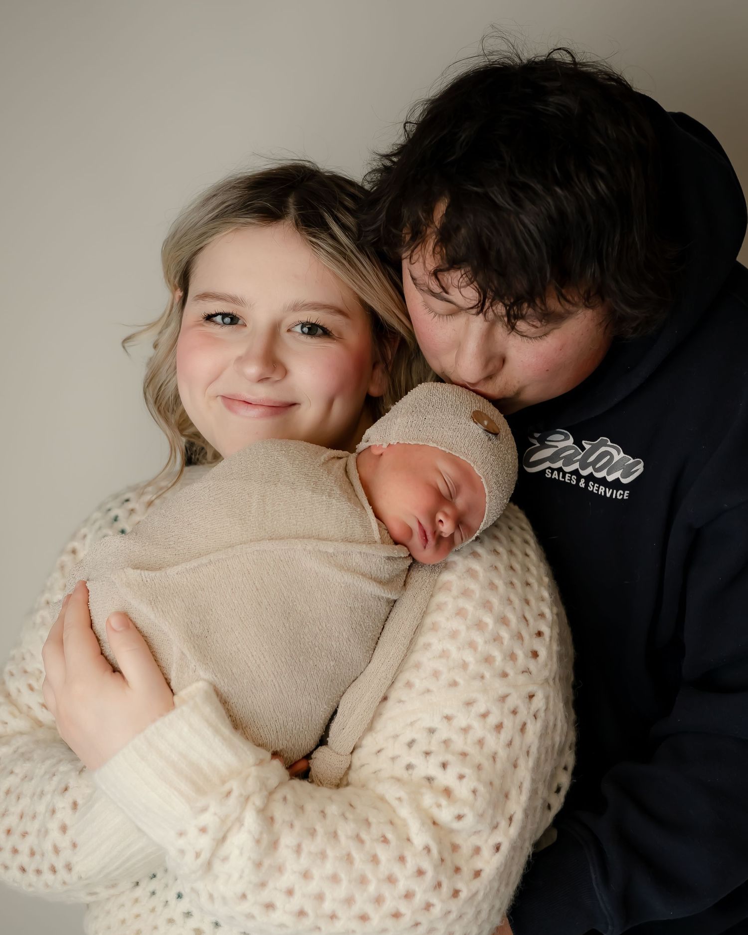 Intimate family portrait of parents holding their sleeping newborn baby wrapped in a beige blanket.