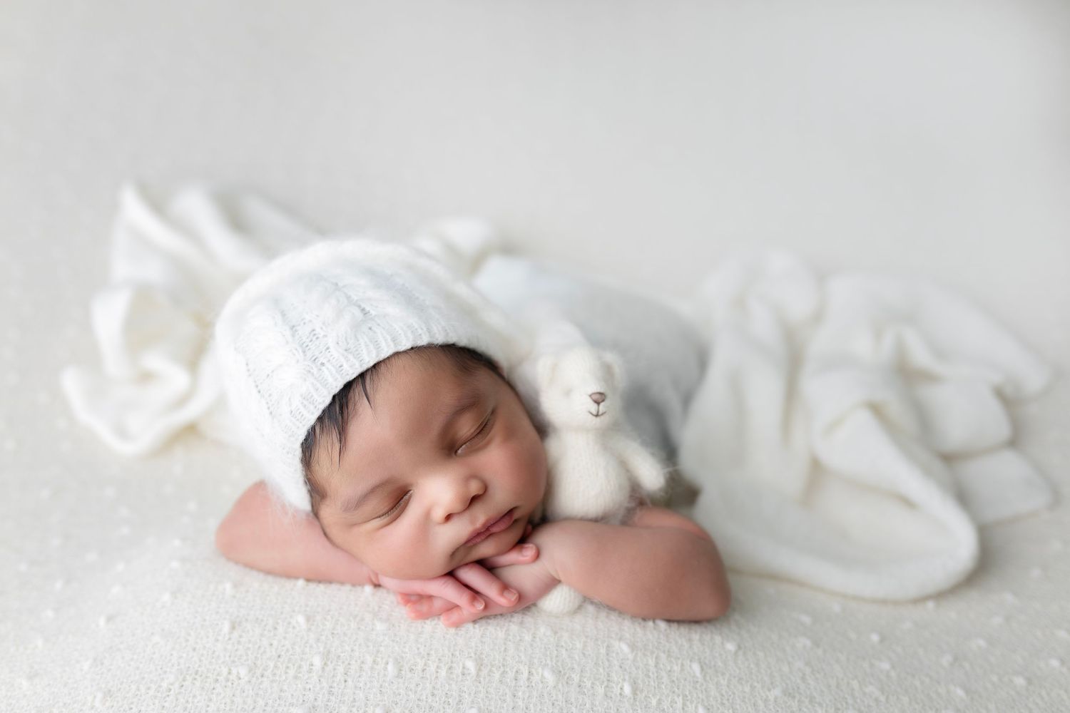 Sleeping newborn wearing white bonnet and wrap against white textured background.