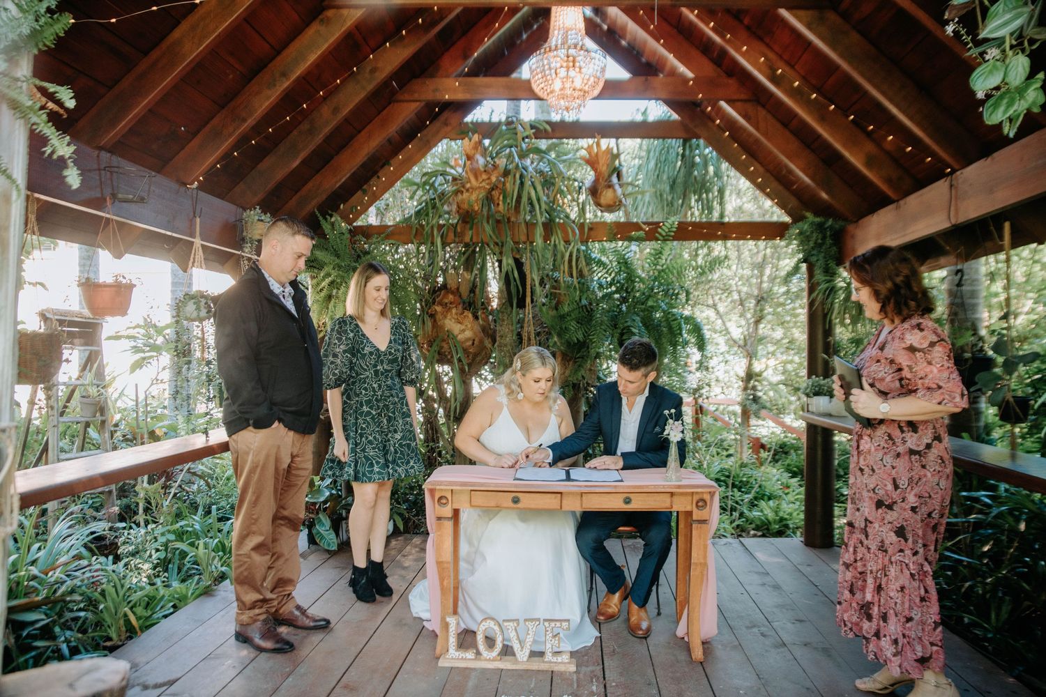 A small wedding ceremony takes place in an intimate wooden pavilion surrounded by tropical plants.