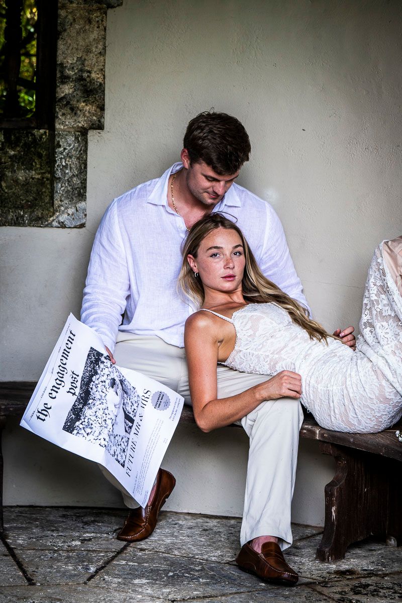 A stylish couple poses together on a bench with a newspaper.