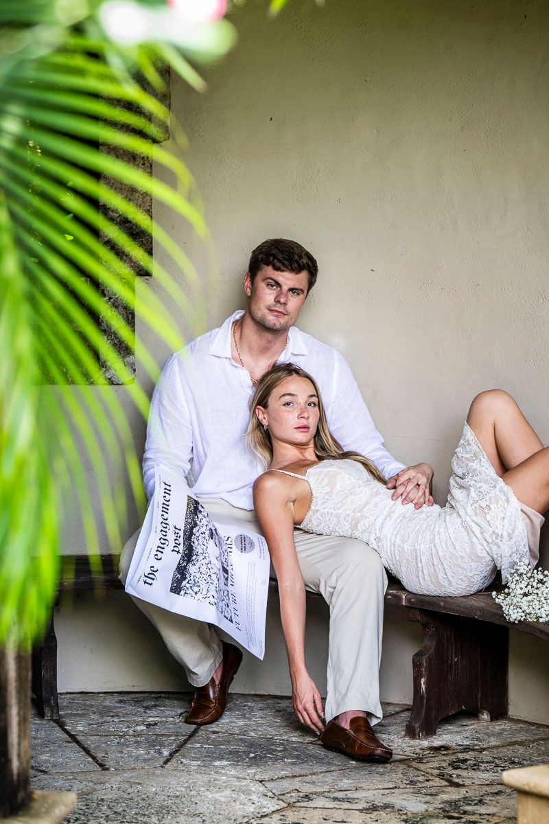 Couple in white attire lounging on bench framed by tropical palm leaves.