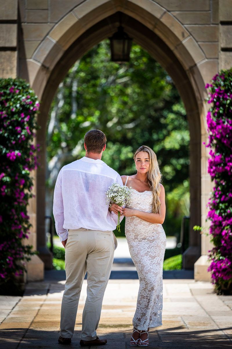 A couple stands beneath a stone archway adorned with vibrant purple flowers during a wedding ceremony.