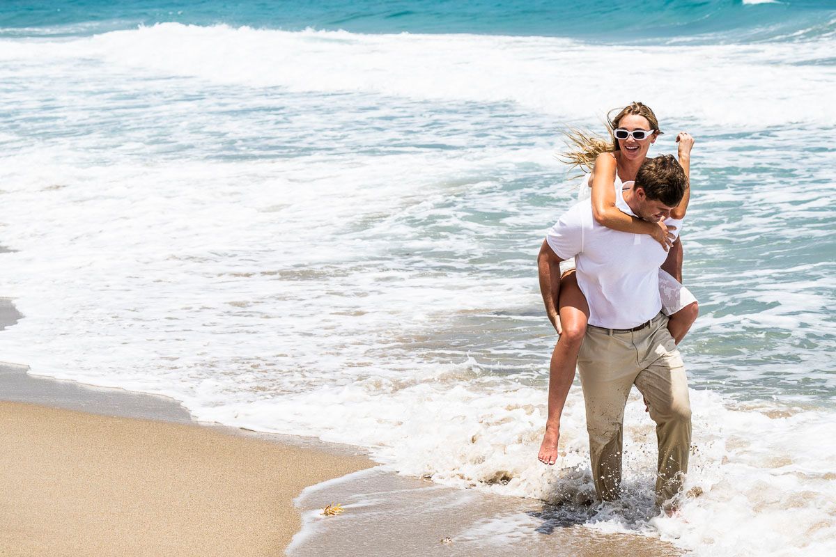 Playful couple enjoying waves at beach shore with white attire.