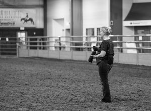 Photographer taking pictures in a dimly lit indoor horse arena with metal railings.