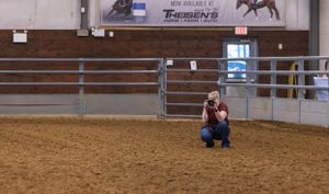 Photographer kneeling in an indoor equestrian arena with dirt floor and metal railings.