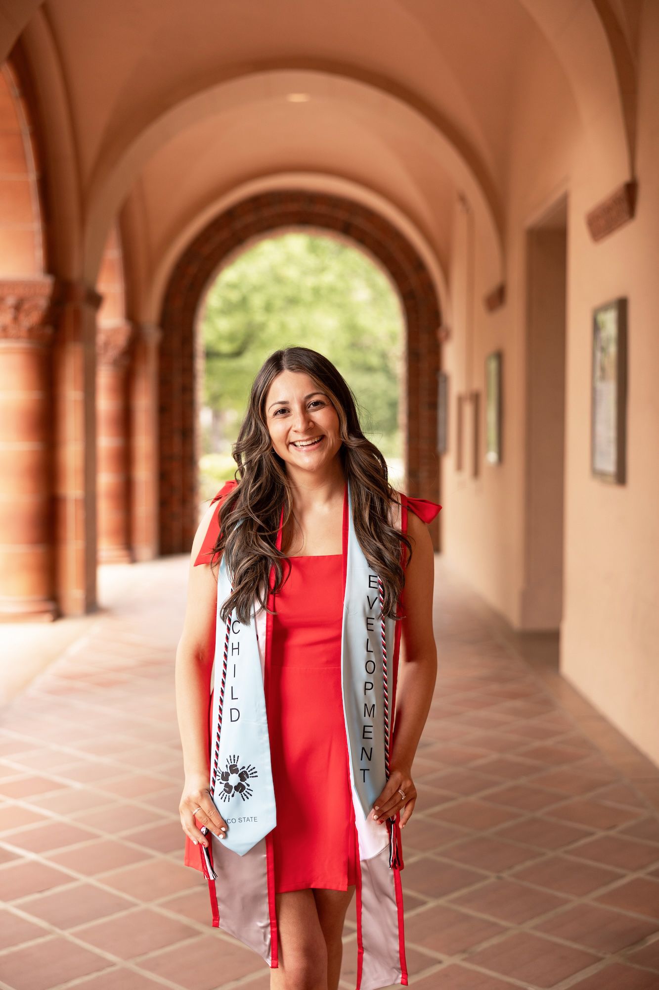 Smiling graduate in red dress wearing sash in arched university hallway.