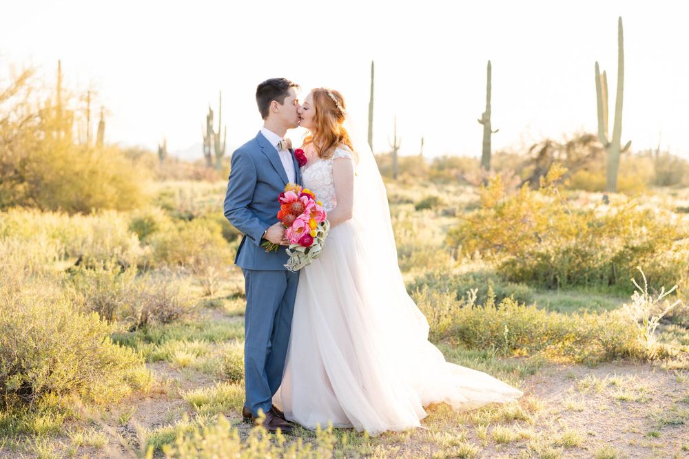 Newlyweds in a desert landscape with saguaro cacti at sunset, bride holding vibrant pink bouquet.