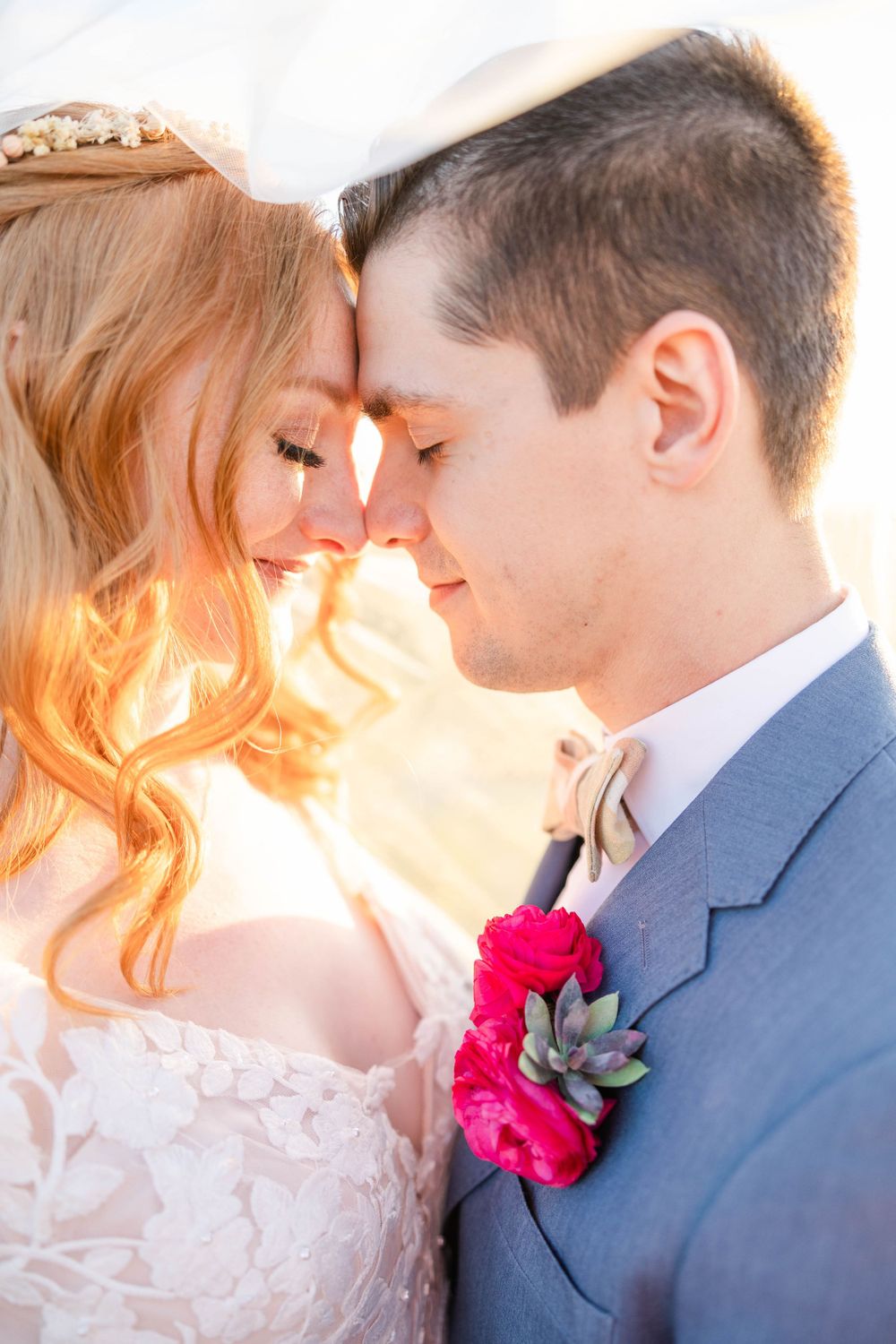 A romantic wedding portrait of the couple touching foreheads with a bright pink floral boutonniere against a sunlit backdrop.