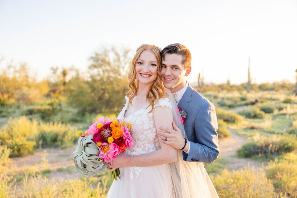 Wedding couple embracing in desert landscape with bright floral bouquet at golden hour.