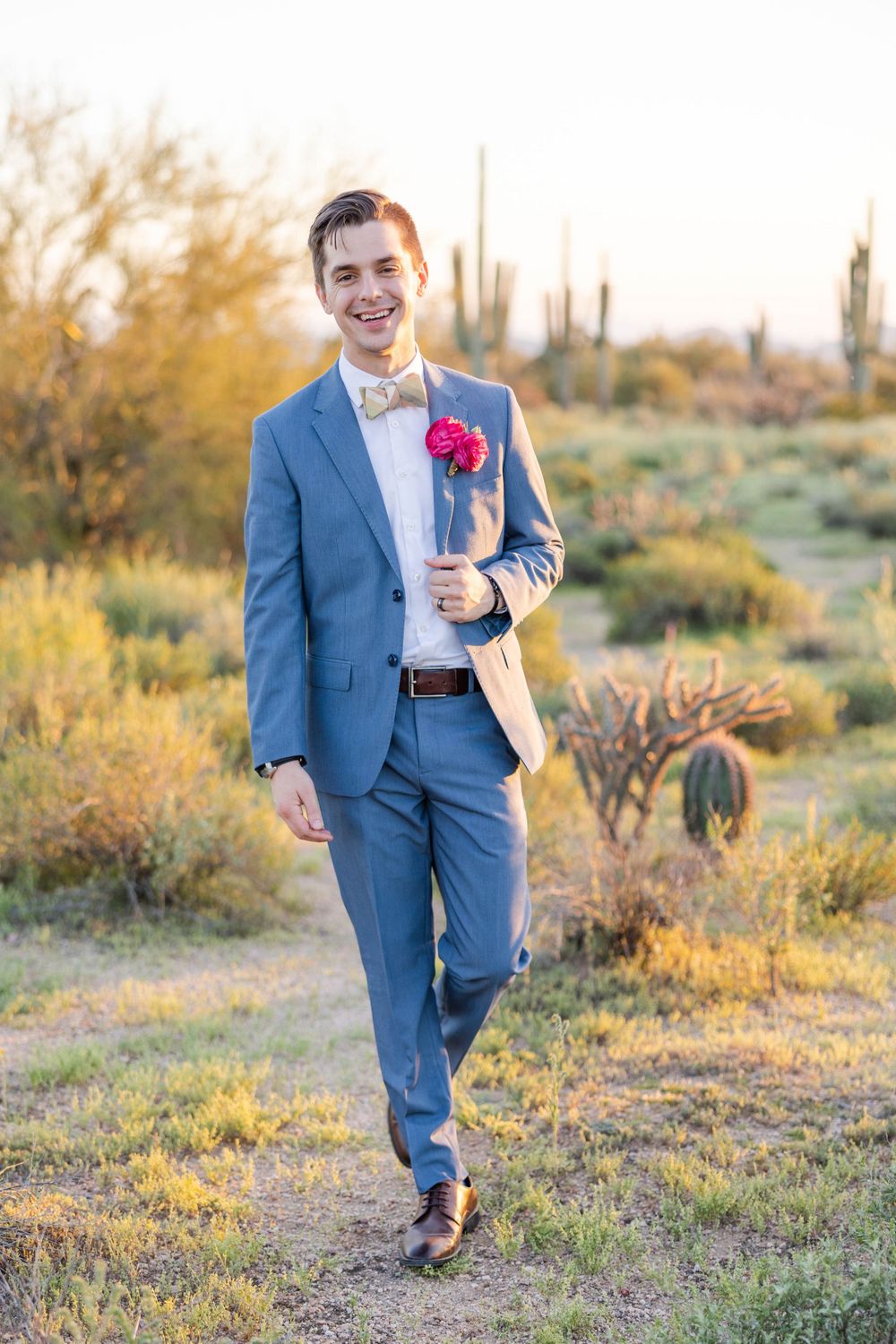 Person in blue suit with pink boutonniere poses in desert landscape with saguaro cacti at sunset.