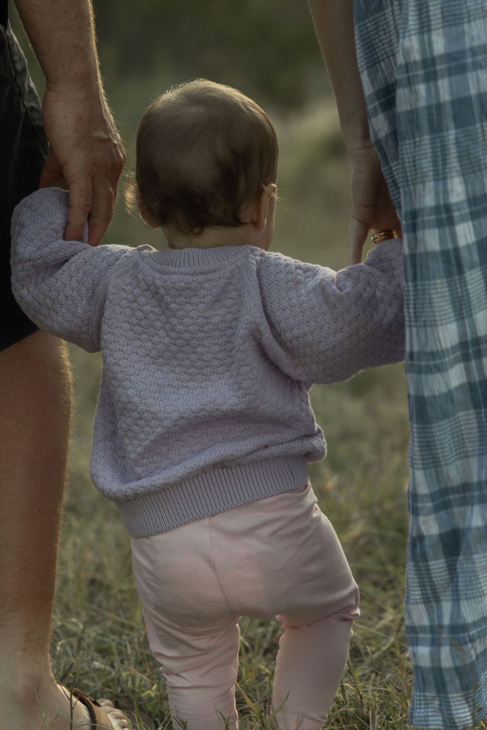Close-up view of a baby walking in grass holding her parents hands during golden hour.