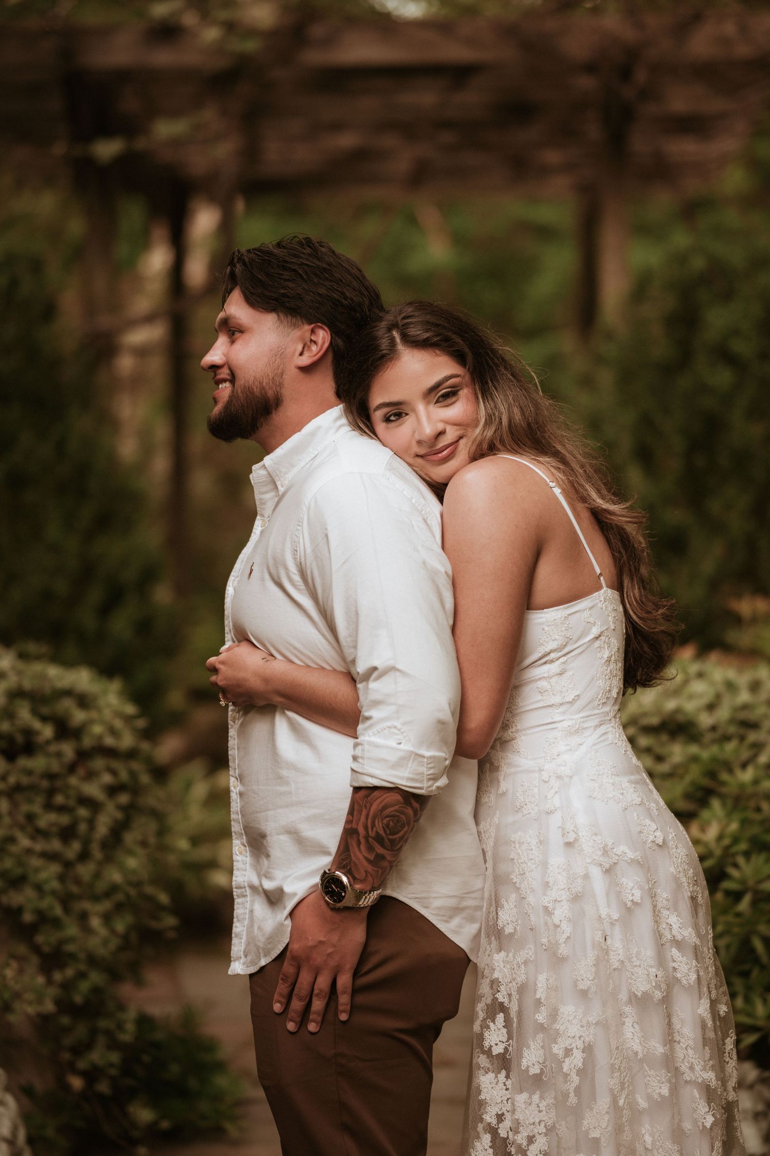 A man in a crisp white shirt faces to the side of the garden while a girl in a white dress hugs him from behind staring ahead with a smile