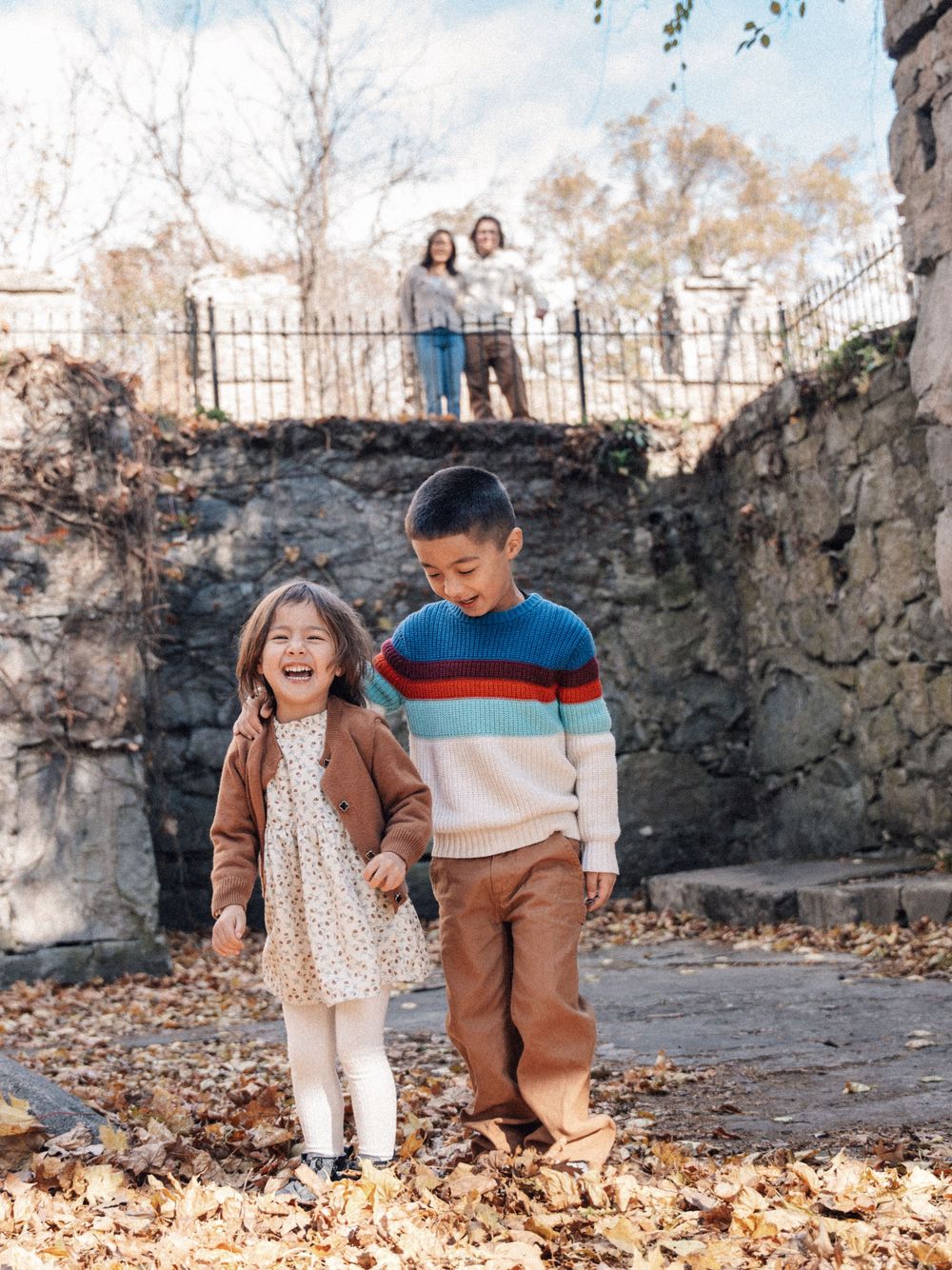 Playful autumn photoshoot with fallen leaves and stone wall backdrop.