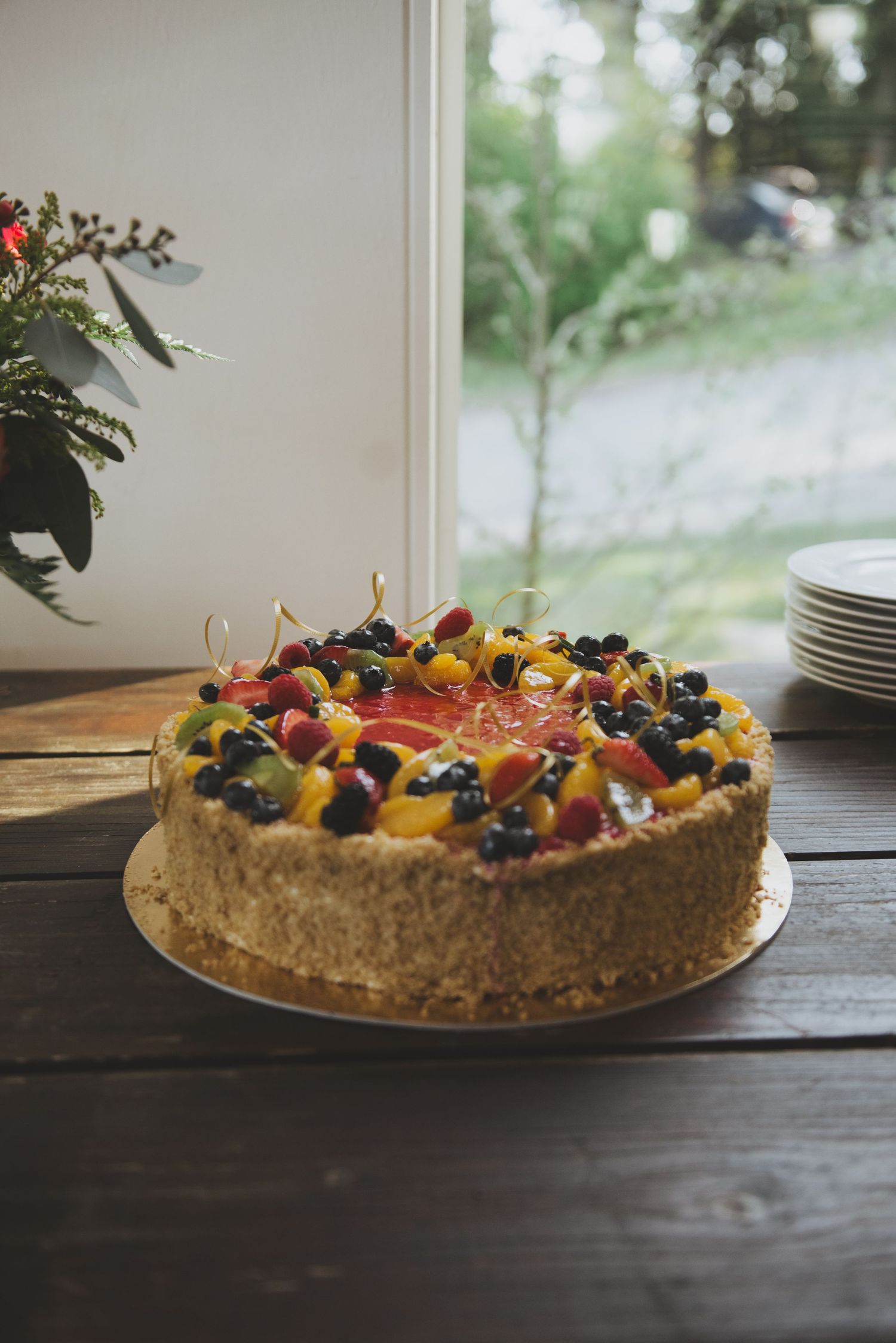 Fresh fruit topped cheesecake on wooden table with white plates and floral arrangement in background.