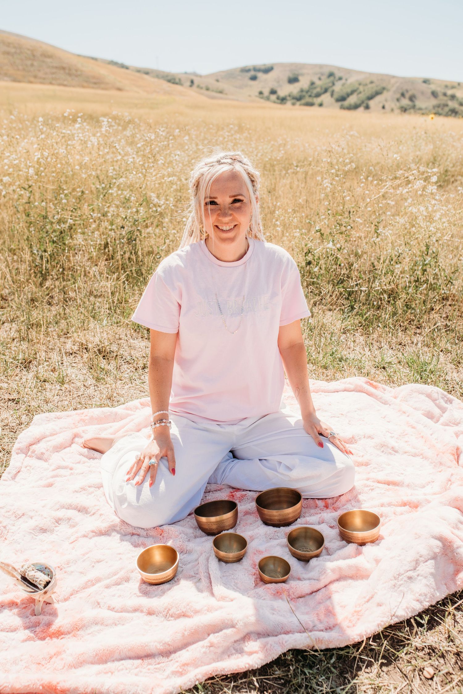 Person in white shirt sitting with singing bowls on pink blanket in sunny meadow.