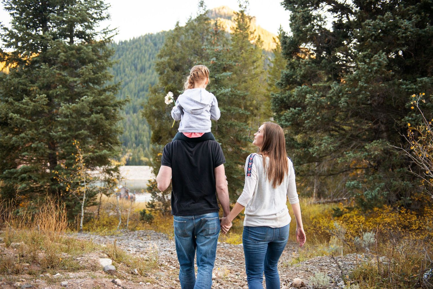 Family walking through mountain trail during golden hour with child riding on shoulders.