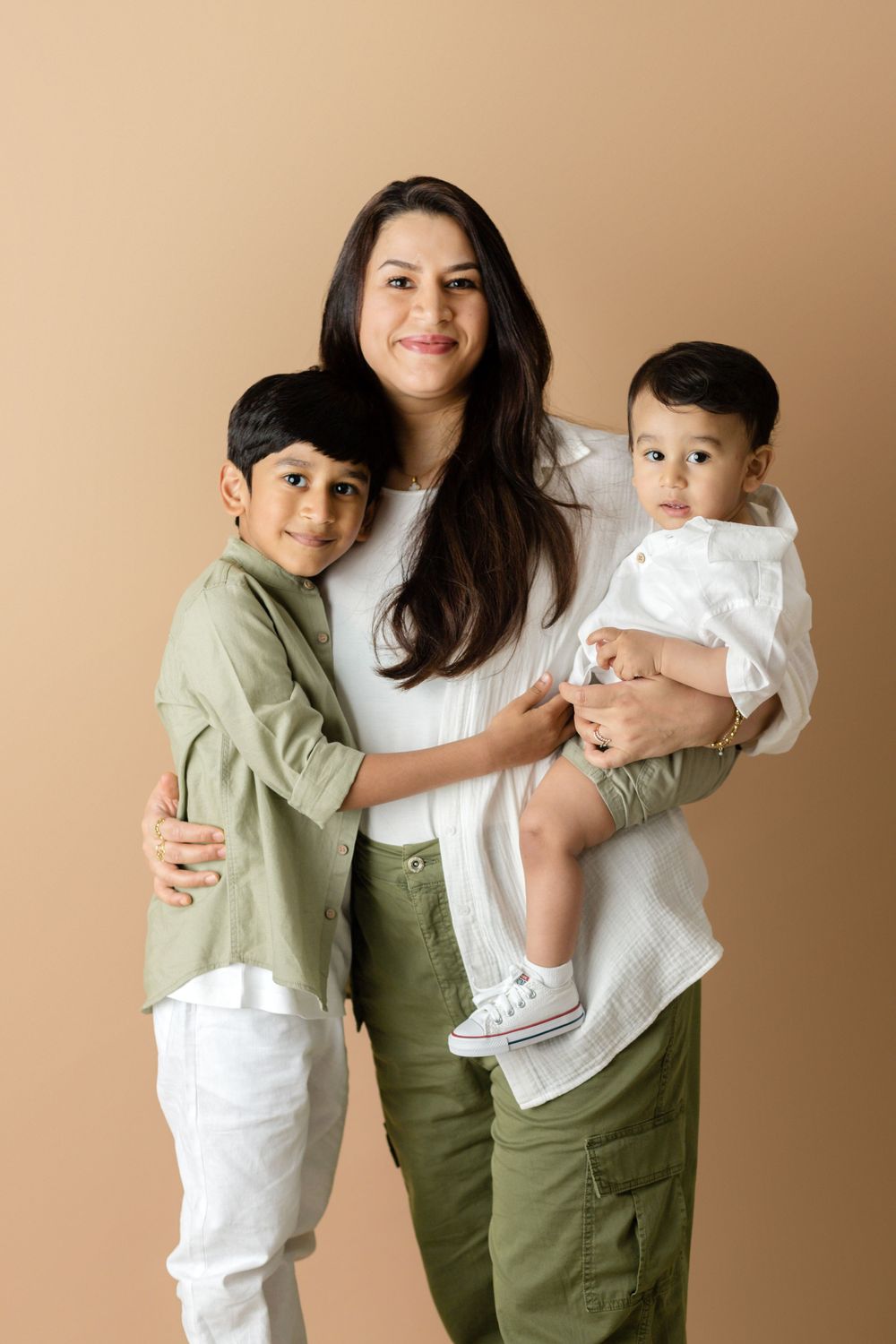 A mother in white top and green pants with two children in casual attire posing against beige wall.